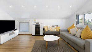 A modern living room with a flat-screen TV, a gray sofa with yellow cushions, a white coffee table, and a large window. - Holiday Home 'Palheiro Dis 3’as' with Sea View, Wi-Fi and Air Conditioning (Achadas da Cruz)