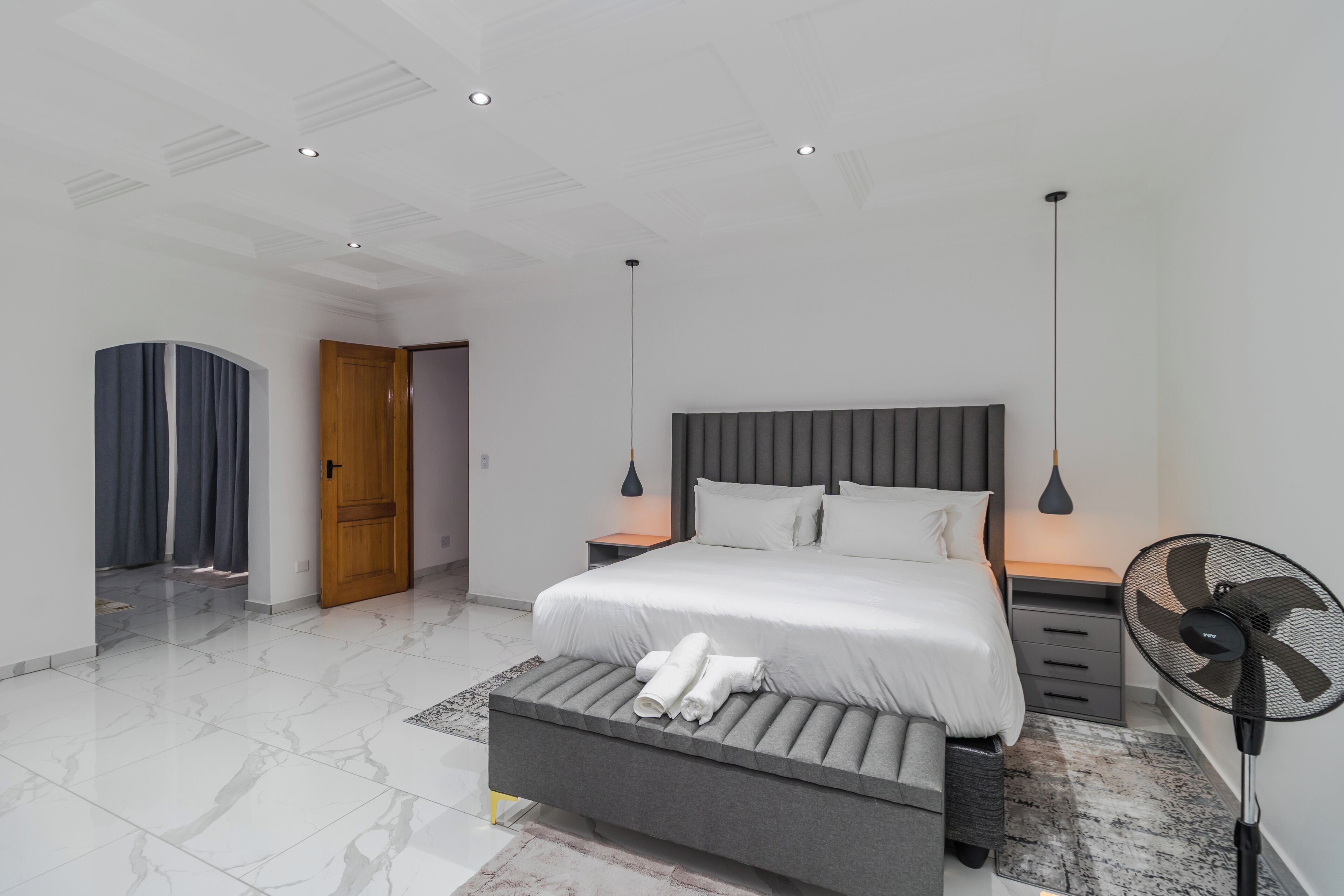A modern bedroom with a bed, bedside tables, a bench, a standing fan, and a door leading to another room.