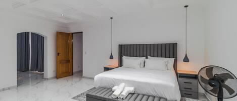 A modern bedroom with a bed, bedside tables, a bench, a standing fan, and a door leading to another room.