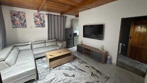 A living room with a sectional sofa, a wooden coffee table, a flat-screen TV, and a kitchen area in the background. - Boutique Hotel-Style Room In Ferndale-G7 (Randburg)