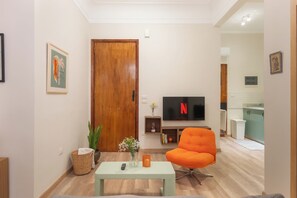 Ein Wohnzimmer mit Holztür, Fernseher, einem leuchtend orangefarbenen Sessel, einem kleinen Tisch und einem Korb.