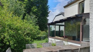 A wooden table and chairs set on a patio with a glass enclosure, surrounded by greenery and a building with large windows. - Cottage, sauna, billiards, 4 bedrooms, 3 bathrooms. Families or friends. (Chiny)