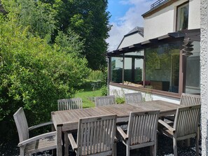 Ein Holztisch mit Stühlen steht auf einer Terrasse mit Glasveranda, umgeben von Grünpflanzen und einem Gebäude mit großen Fenstern.