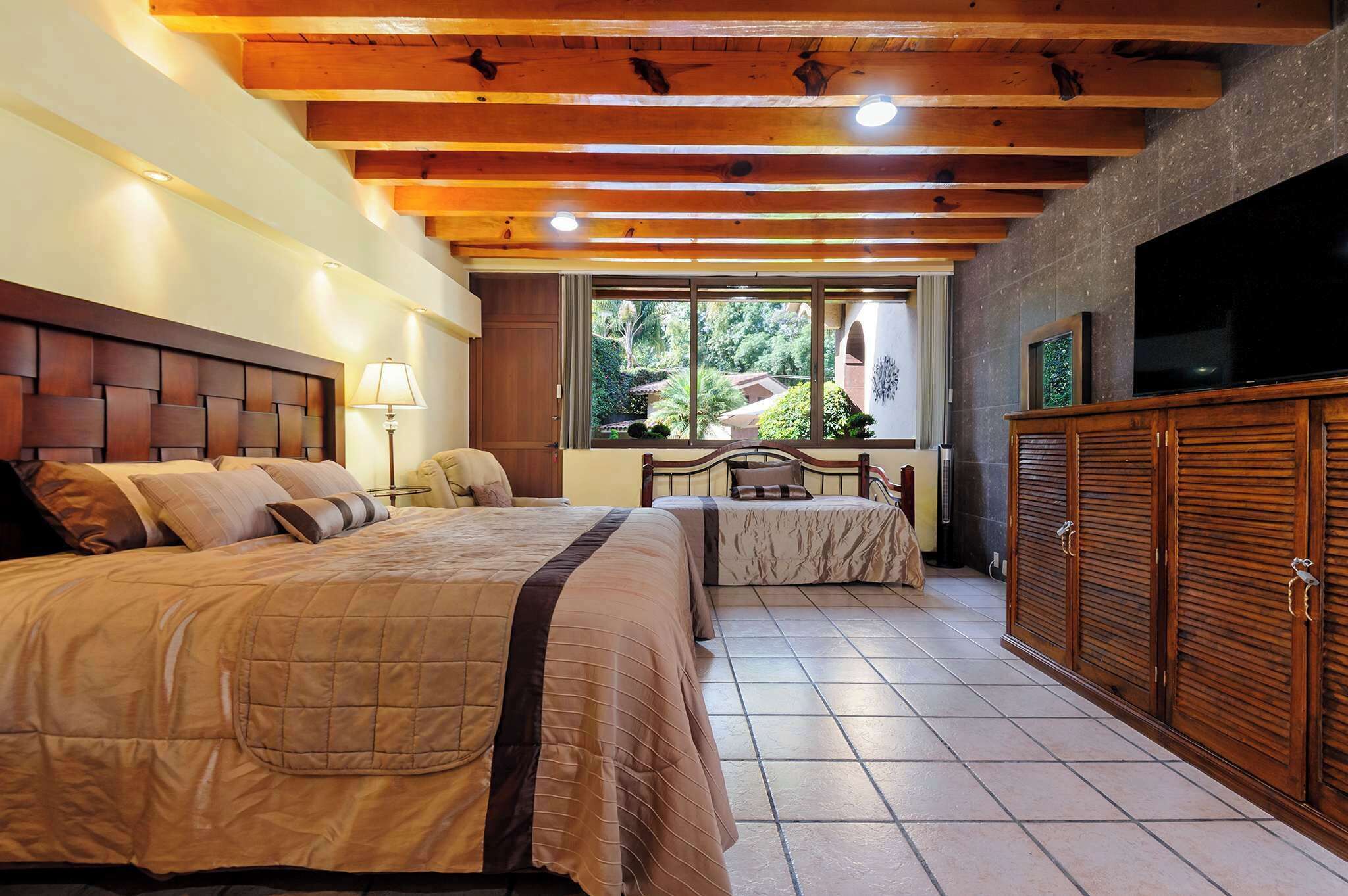 A bedroom with two beds, a TV, a wardrobe, and a window with a view of greenery.