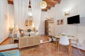 A bedroom with a bed, bedside table, curtains, a TV, and a dining area with chairs and a table. - Le Caravelle - Apt 38 Apartment (Castellammare del Golfo)