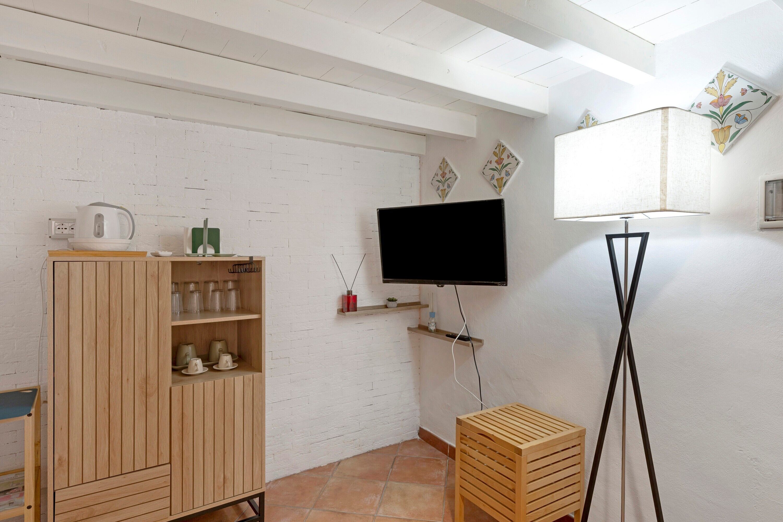 Ein Zimmer mit weißer Ziegelwand, einem Holzkabinett mit Regalen, einem wandmontierten Flachbildfernseher, einer Stehlampe und einem kleinen Holztisch.