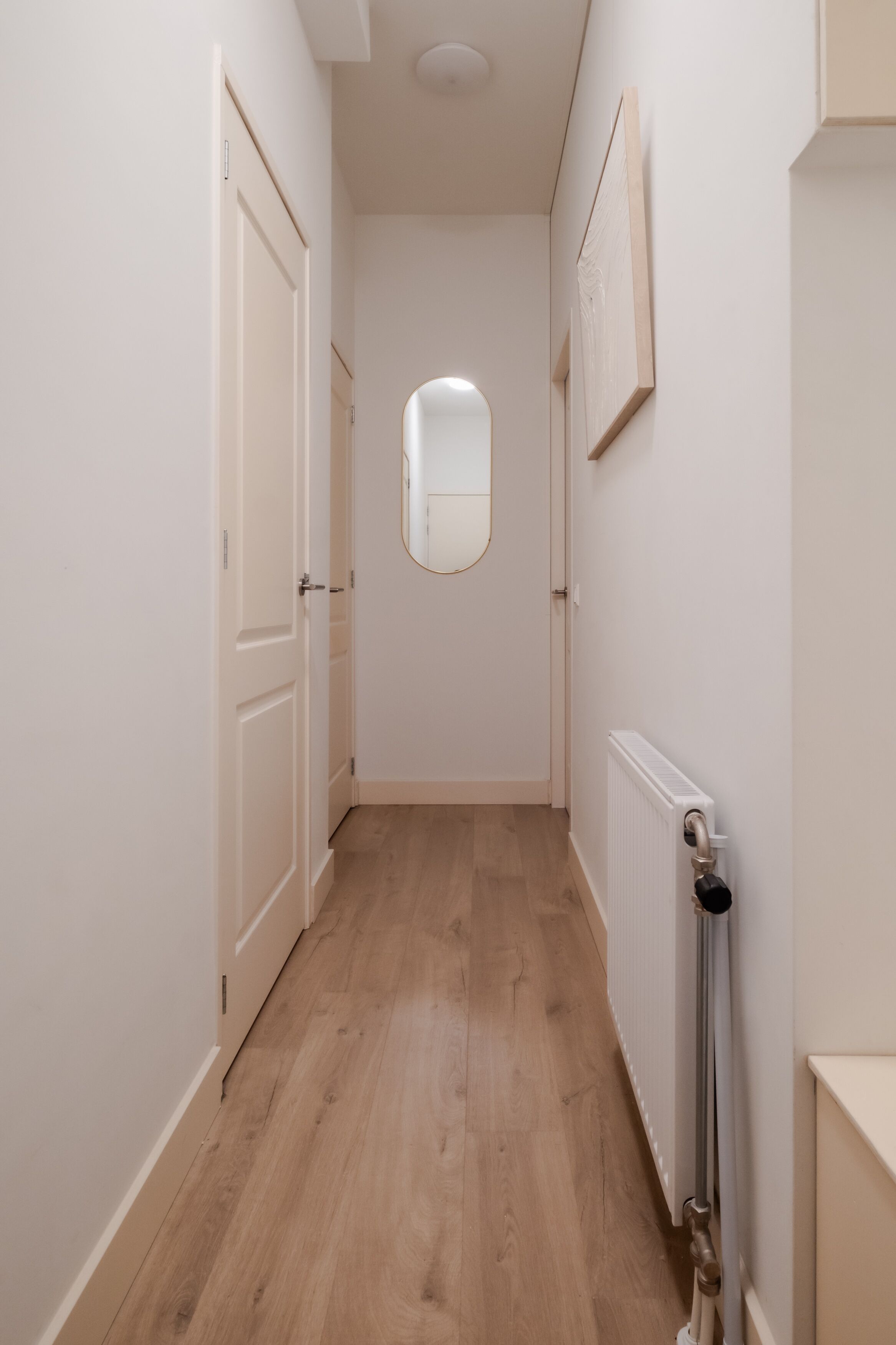 A narrow hallway with wooden flooring, white walls, a radiator, and a door on the left.