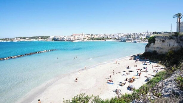 Een kuststrand met helder water, een rotsachtige klif en een stadsgezicht op de achtergrond.