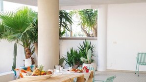 Een houten tafel met een fruitschaal, groene stoelen en potplanten in een lichte kamer met witte muren en een grote kolom.