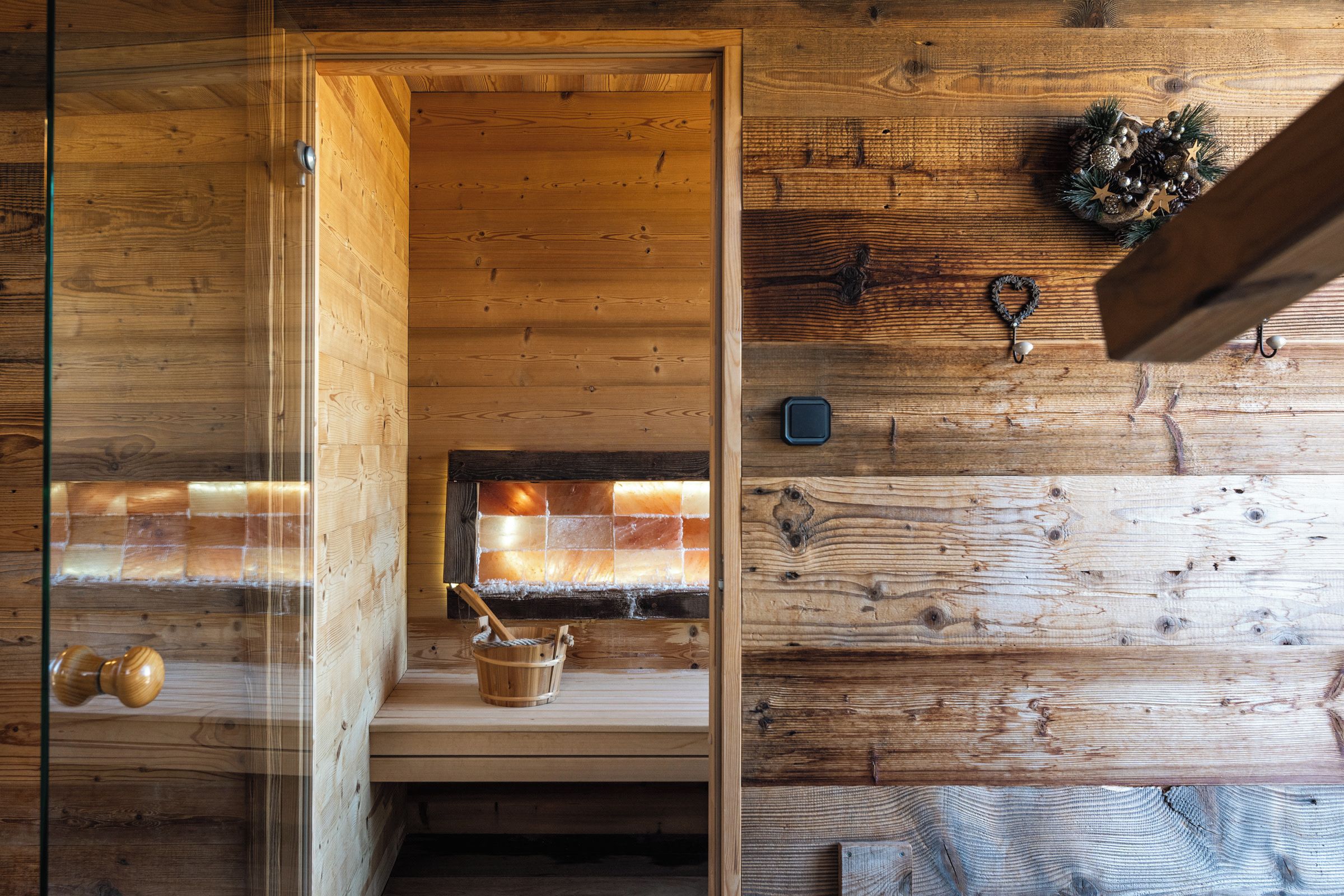 Interior de una cabaña de madera con un ventanal que da a una chimenea, un banco de madera y una pared rústica con ganchos.