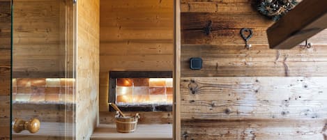 Interior de una cabaña de madera con un ventanal que da a una chimenea, un banco de madera y una pared rústica con ganchos.