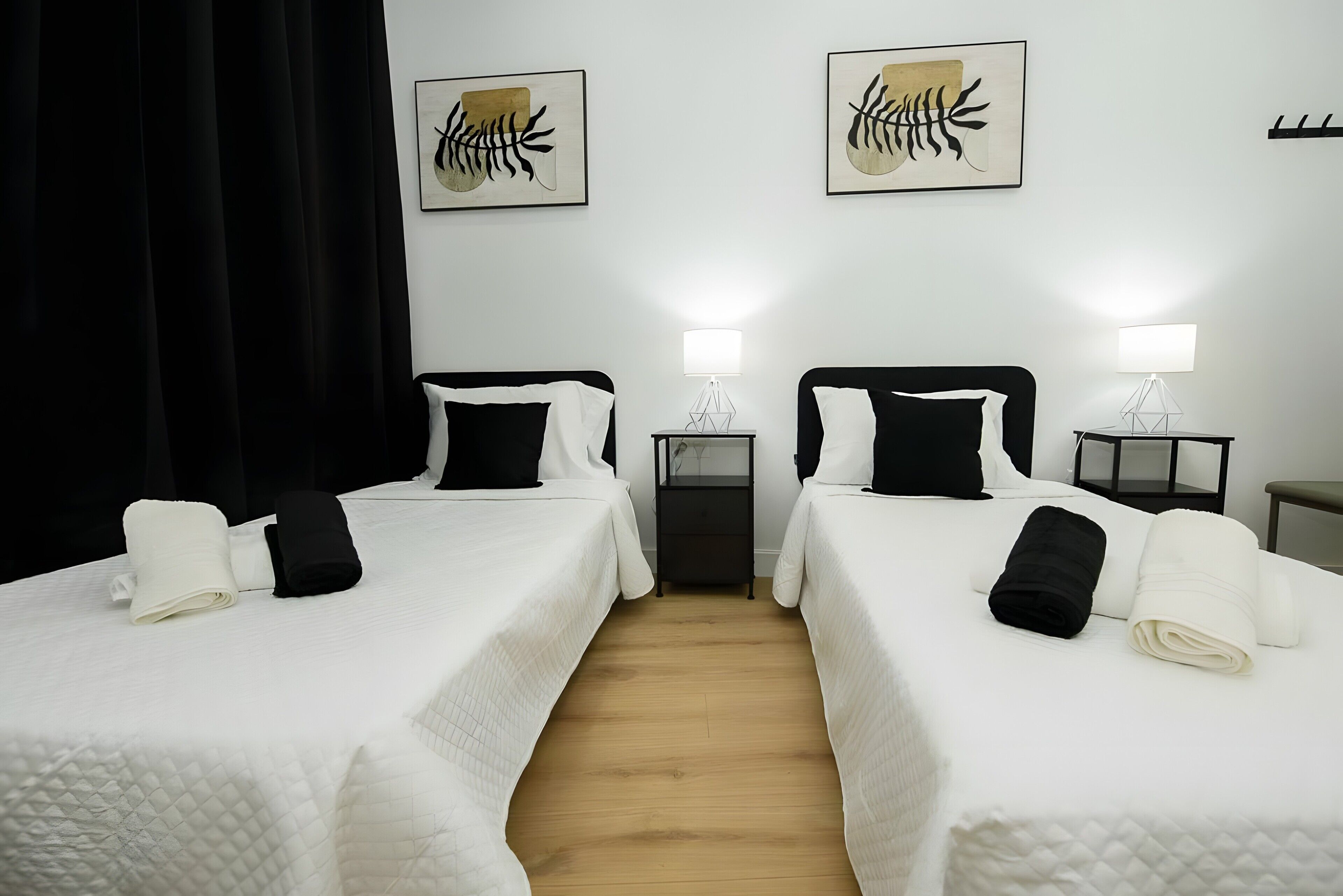 Habitación de hotel con dos camas, ropa blanca, almohadas negras y piso de madera.