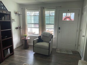 Ein Wohnzimmer mit einer Couch, einem kleinen Tisch mit einer Pflanze, einem Bücherregal und einer Tür mit Fußmatte.