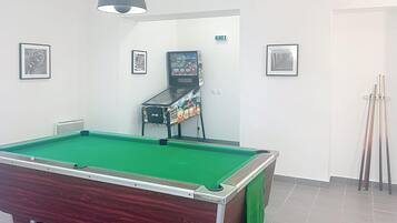 A pool table with a green felt surface, a wooden table base, and a green cloth draped over one side. There is a small, enclosed arcade machine in the background, and a pair of pool cues leaning against the wall.