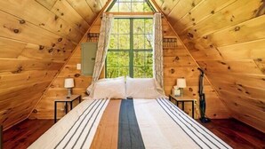 Quarto de sótão com madeira, uma cama, duas mesas de cabeceira, uma janela com cortinas e vista para o verde lá fora.