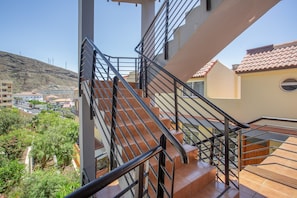 Un escalier avec des rampes en métal mène à un balcon qui donne sur un quartier résidentiel, avec des maisons et une colline en arrière-plan.
