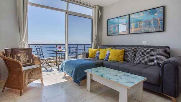 Uma varanda com vista para o mar, um sofá com almofadas, uma cadeira de vime e uma mesa com tampo de vidro.