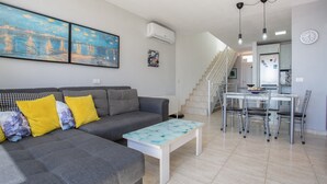 Uma sala de estar com um sofá cinzento, almofadas amarelas, uma mesa de centro branca e uma escada que leva a uma área de cozinha.