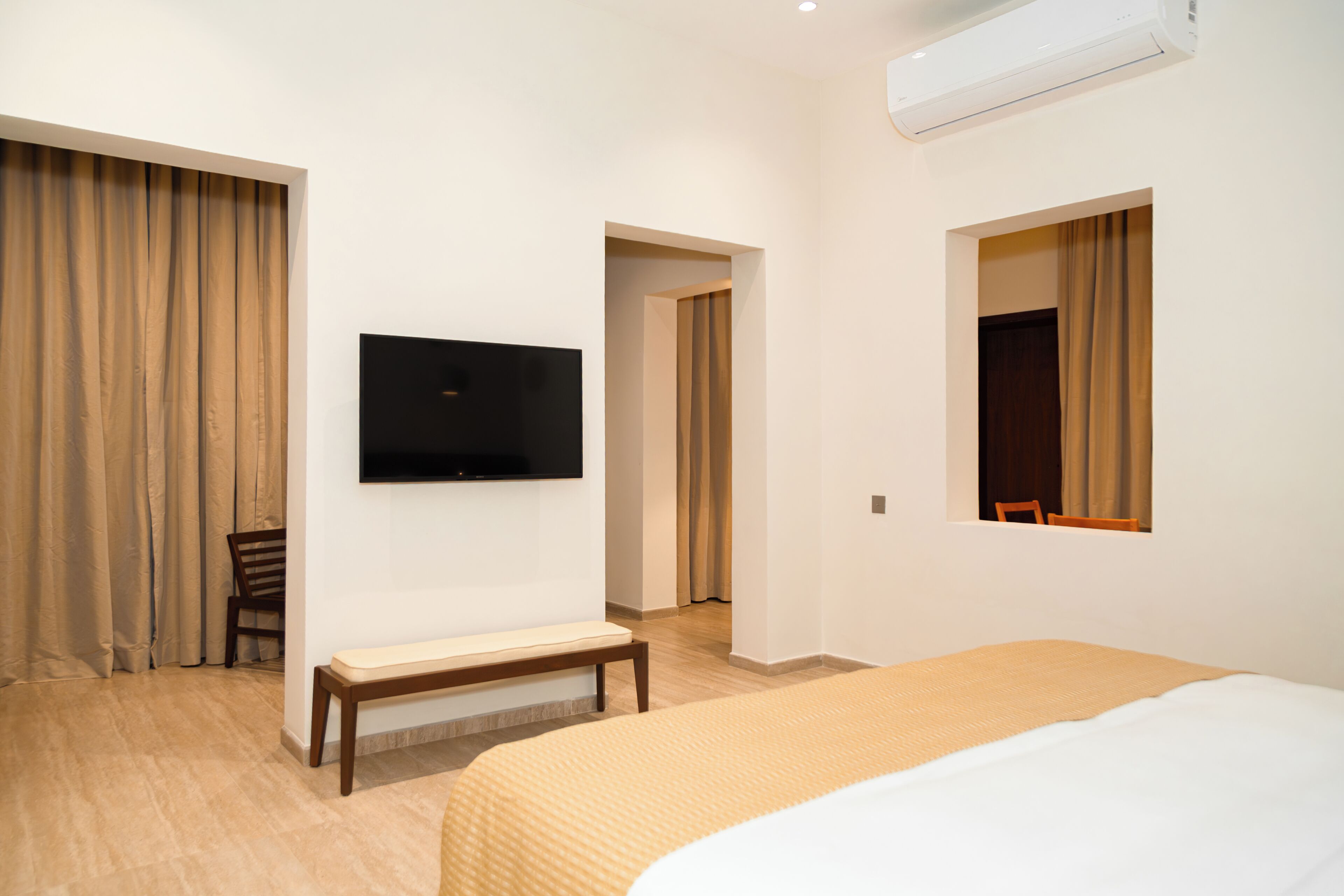 A hotel room with a bed, a TV mounted on the wall, a bench, and a window with curtains.