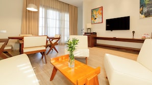 Habitación de hotel con sofá, mesa de centro de madera con un florero, mesa de comedor con sillas, televisor de pantalla plana y un cuadro en la pared.