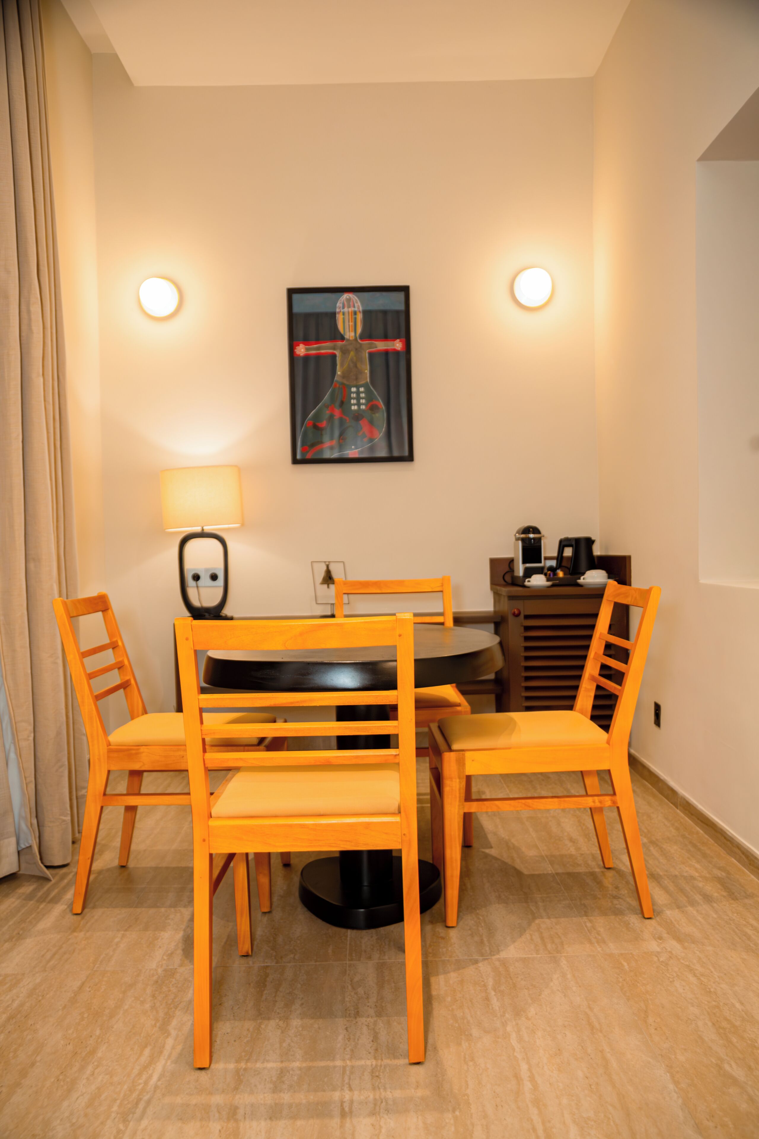 Uma área de refeições com uma mesa redonda, quatro cadeiras, uma luminária, um quadro emoldurado e uma cafeteira.