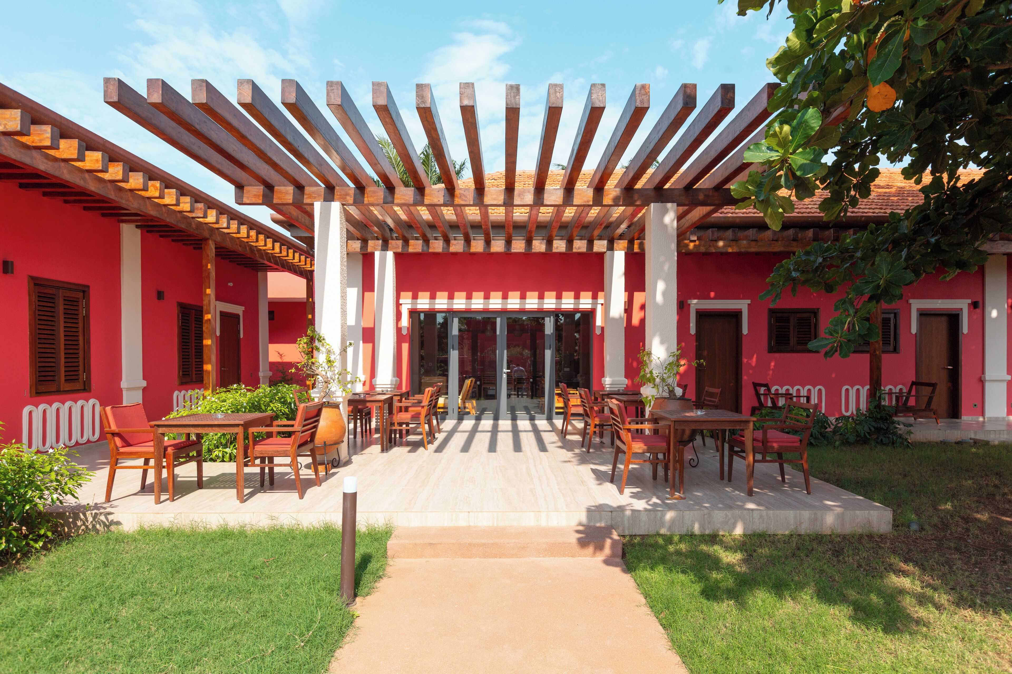 Un área de estar al aire libre con techado, mesas y sillas, un camino que conduce a la entrada de un edificio y una fachada roja.
