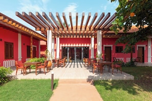Un área de estar al aire libre con techado, mesas y sillas, un camino que conduce a la entrada de un edificio y una fachada roja.