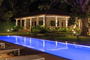 A pool with a blue light, a covered outdoor area with steps, and a large tree. - Native Hotels (Ouidah)