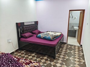 Quarto com cama, mesa de cabeceira, tomada na parede e banheiro visível por uma porta aberta.