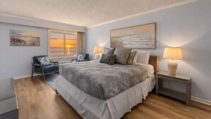 Een slaapkamer met een groot bed, een stoel, een nachtkastje en een raam met uitzicht op een zonsondergang.