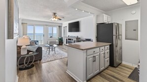 Een moderne keuken met een centraal eiland, een eethoek met tafel en stoelen, en een zithoek met een bank en een lamp.