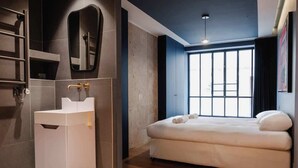 Ein modernes Hotelzimmer mit großem Fenster, einem weißen Bett, einem integrierten Waschbecken und einem Spiegel.