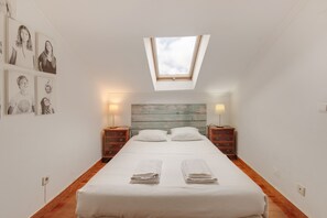 Un dormitorio con tragaluz, una cama con dos almohadas, mesitas de noche con lámparas y una pared con cuadros enmarcados.