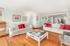 Een woonkamer met twee witte banken, een salontafel met boeken, houten vloeren en ingelijste foto's aan de muur.