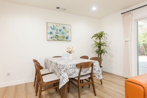 Een eetgedeelte met een tafel voor vier personen, omringd door houten stoelen, een potplant en een lijst met een bloemistisch schilderij aan de muur.