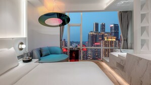A modern hotel room with a large bed, a blue armchair, a round table with a vase, and a cityscape view through large windows. - ASPERA HIGH-ALTITUDE GARDEN PANORAMIC (Chongqing)