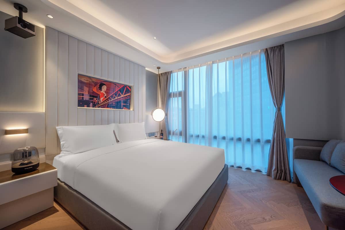 A modern hotel room with a large bed, a bedside table with a lamp, a sofa, and a large window with curtains.