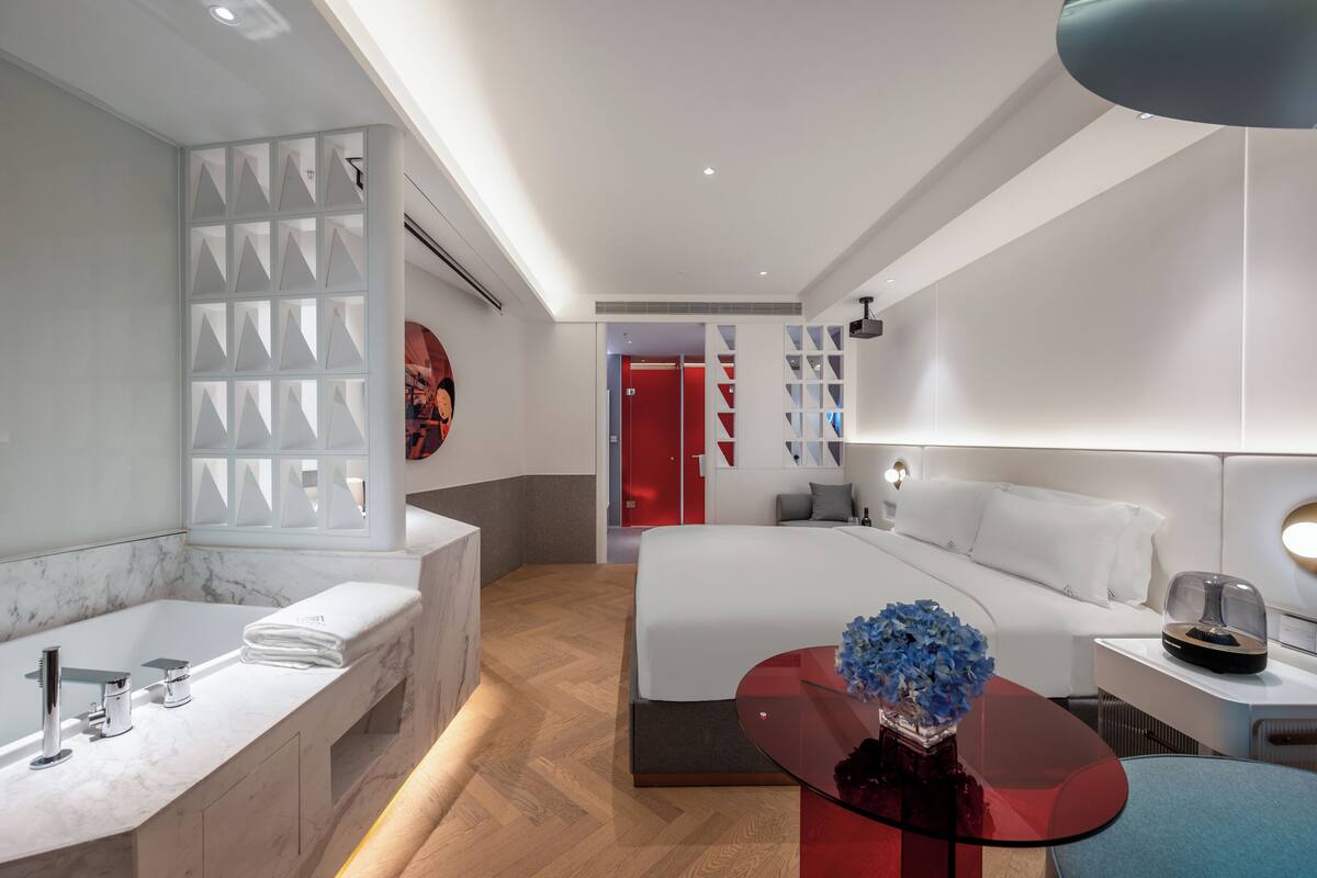 A modern hotel room with a marble bathroom, a glass coffee table with flowers, a bed with white bedding, and a red accent wall.