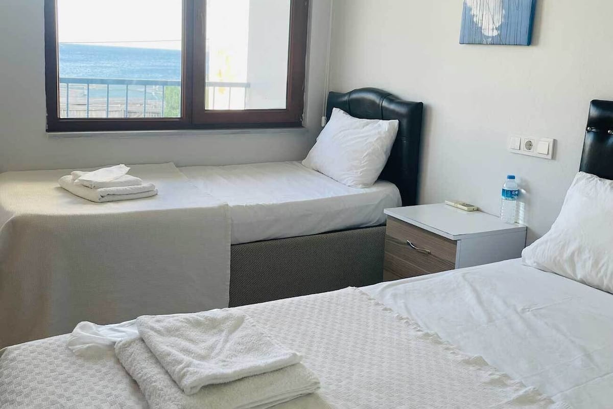 Habitación de hotel con dos camas, una ventana que ofrece vistas al mar, una mesita de noche con una botella de agua y un cuadro en la pared.