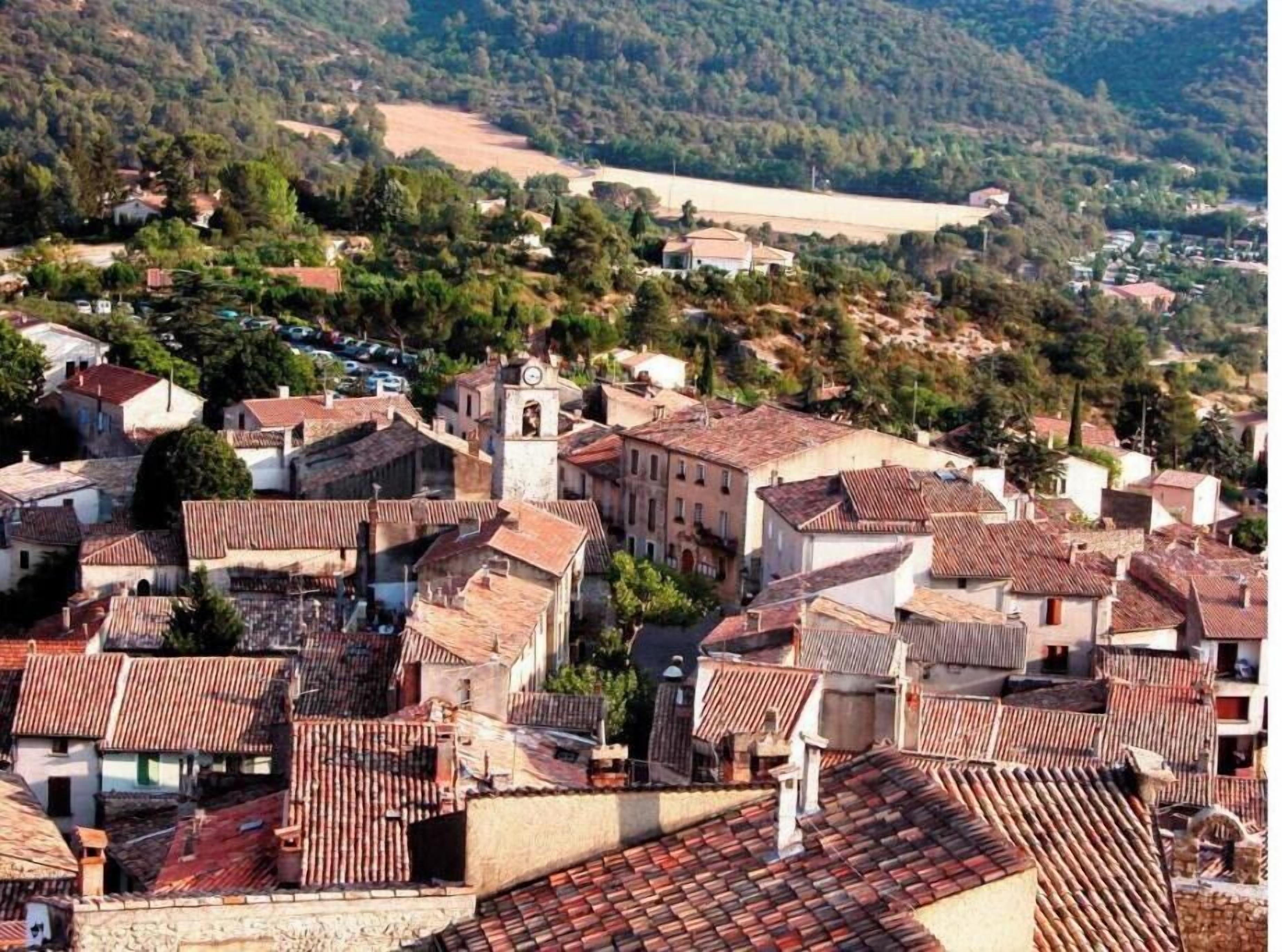 Uma cidade na encosta de uma colina, com edifícios de telhados em terracota, uma igreja com um campanário, e uma estrada que serpenteia a colina.
