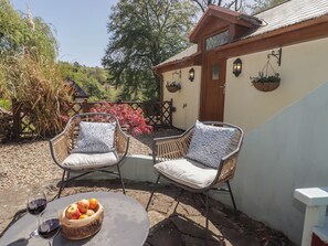 Un patio con sillas de mimbre, una mesa con frutero y copas de vino, y un edificio con puerta de madera y macetas colgantes.