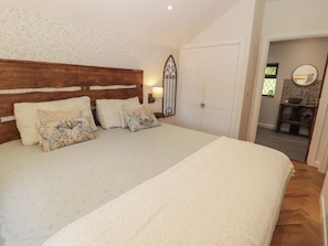 Une chambre à coucher avec une tête de lit en bois, un grand lit recouvert d’une literie à motifs, un luminaire fixé au mur, une armoire blanche et une salle de bain visible à travers une porte ouverte.