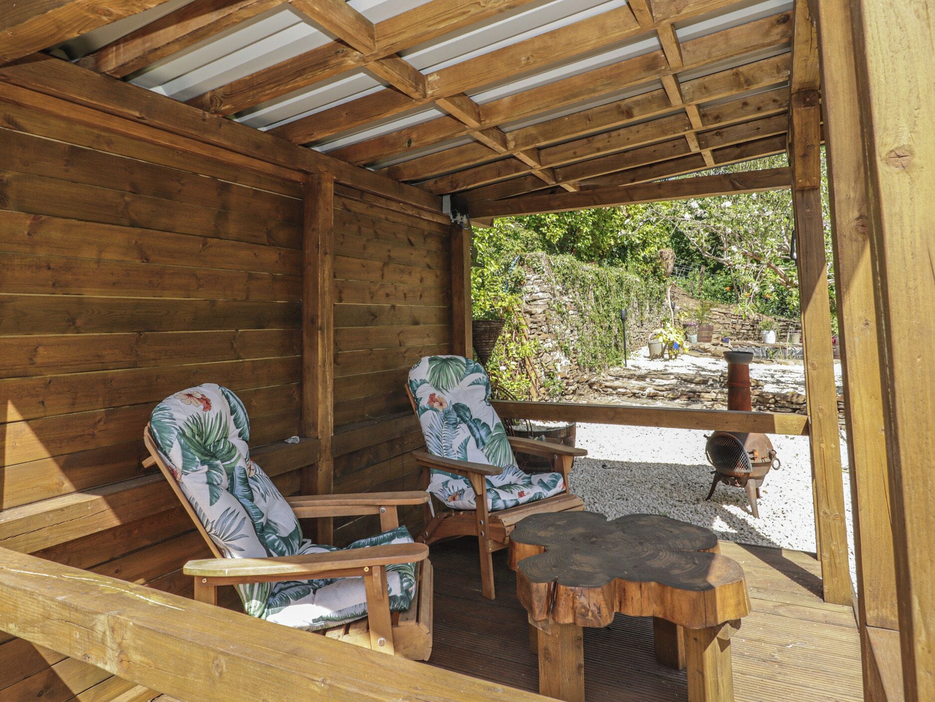 A wooden deck with two chairs and a table, a stone wall, and a view of greenery.