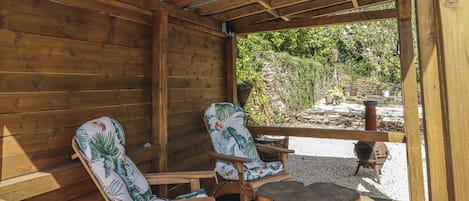 A wooden deck with two chairs and a table, a stone wall, and a view of greenery.