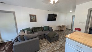 A modern living room with a sectional sofa, a small table, a TV mounted on the wall, and a kitchenette area. - Stargazer Hideaway: Hot Tub, Outdoor Dinning, pet-friendly; close to Yellowstone (Island Park)