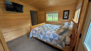 A wooden cabin bedroom with a bed, a television, a window, and a door. - Stargazer Hideaway: Hot Tub, Outdoor Dinning, pet-friendly; close to Yellowstone (Island Park)