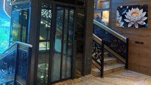 A hotel lobby with a glass elevator, a staircase with ornate railings, and a large floral painting on the wall.
