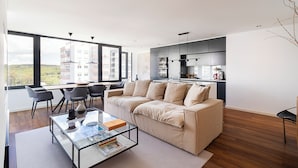Un salon moderne comprenant un canapé, une table basse et des fauteuils, un coin cuisine et de grandes fenêtres offrant une vue sur la ville.