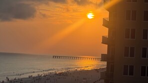 A beach with a pier extending into the water, a building with balconies, and the sun setting in the background.