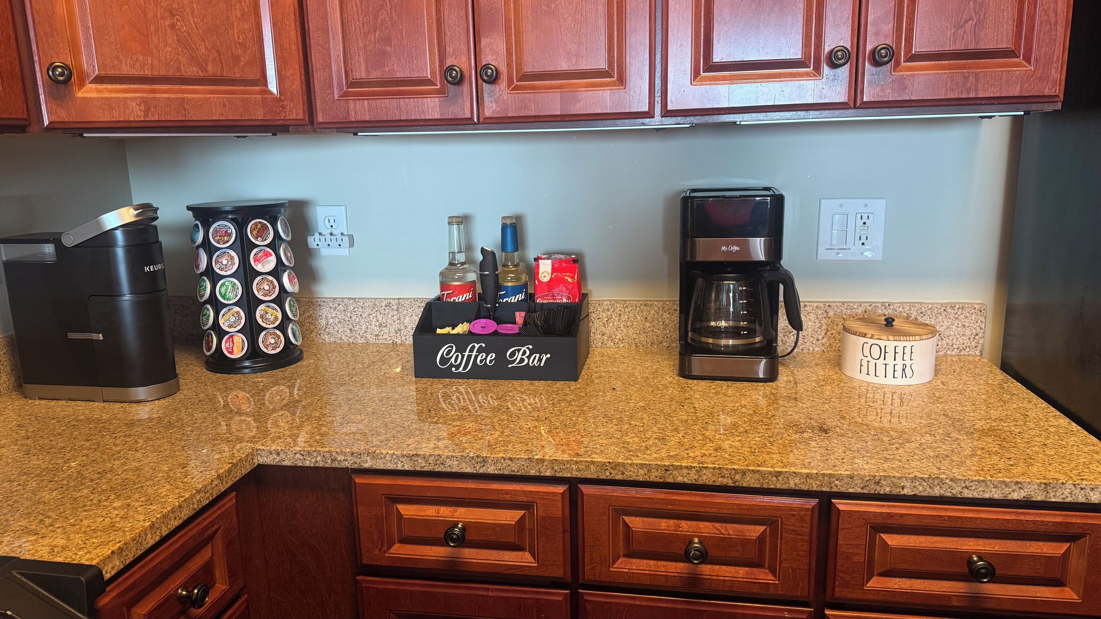 Una encimera de cocina con una cafetera Keurig, un portacapsulas de café, una barra para café, una cafetera Mr. Coffee y un contenedor para filtros de café.